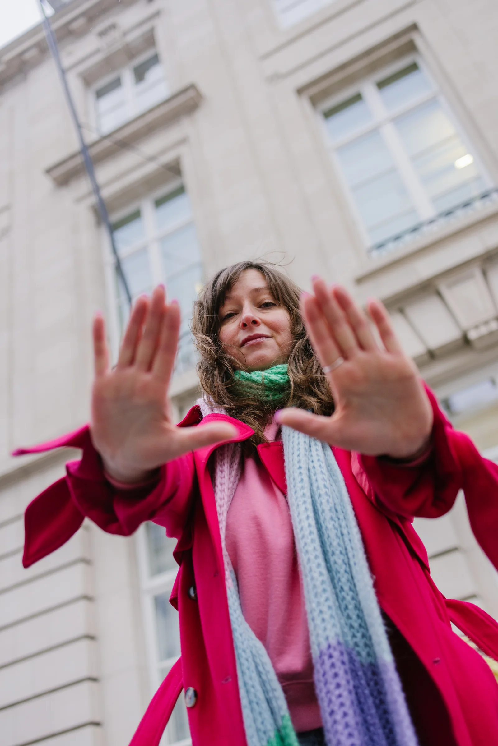 Amélie Beerens in a red coat, both palms raised towards the camera, in front of a Brussels ministry.