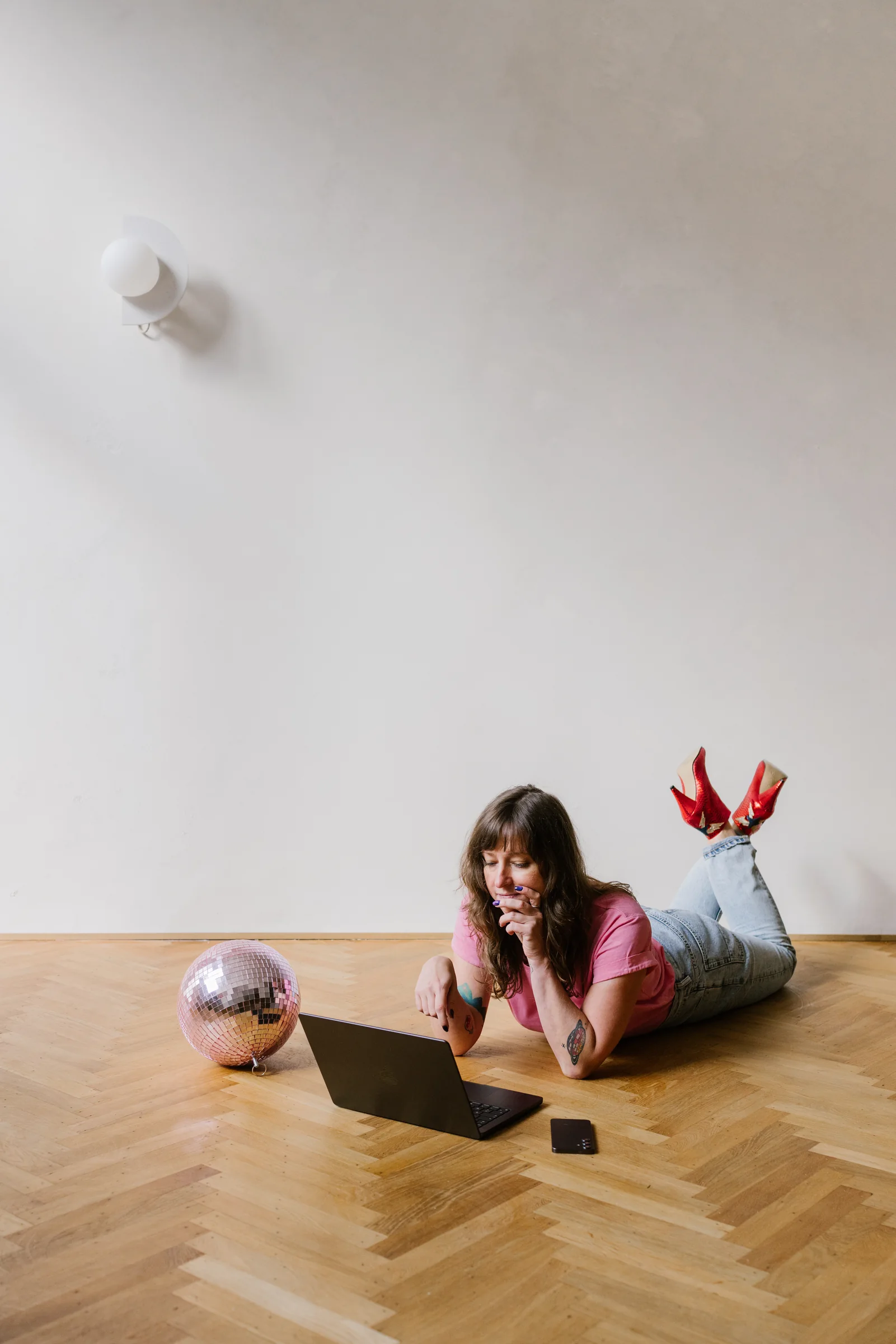 Amélie Beerens lying on a parquet floor, reading on a laptop, a pink disco ball beside her.